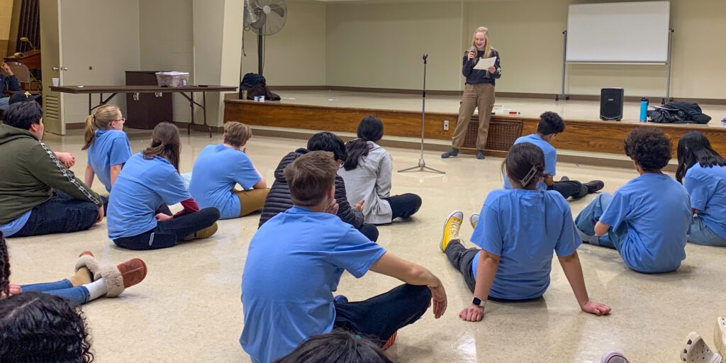 teens sitting on floor and listening to a person who is standing and speaking